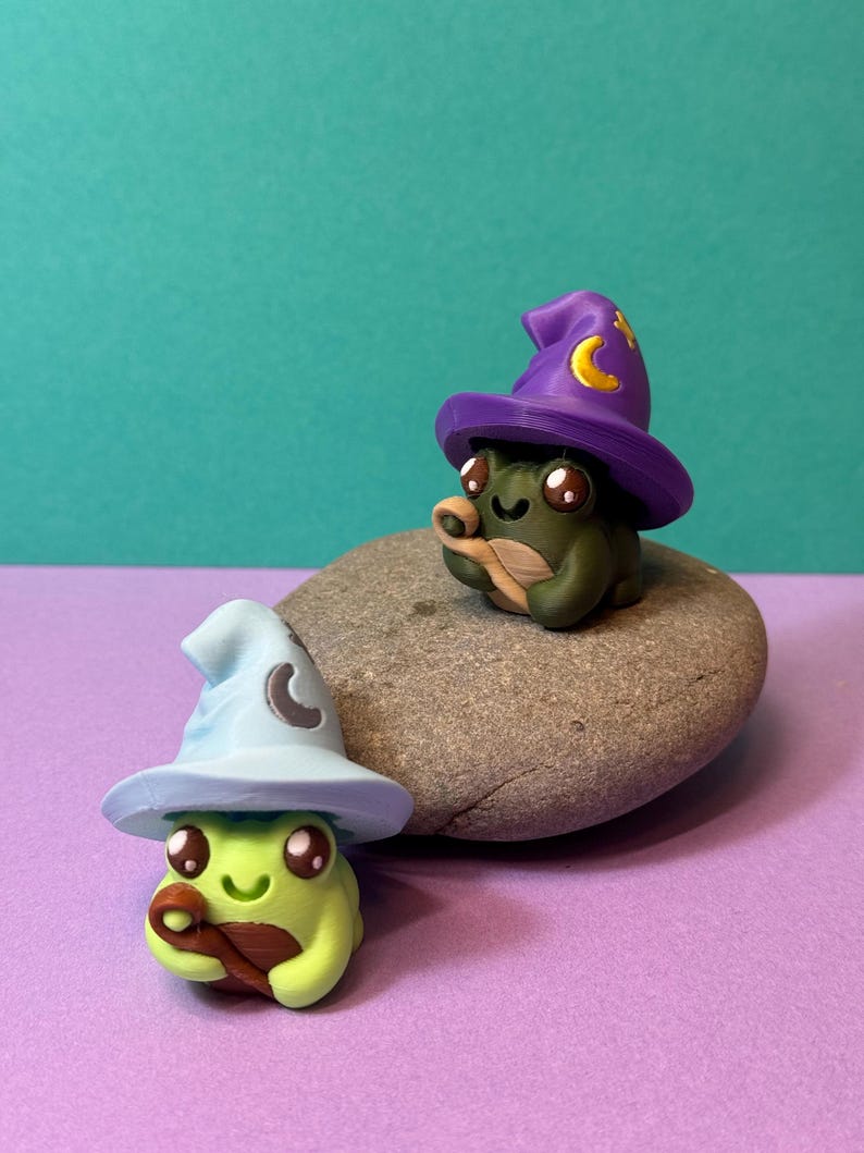 May include: Two whimsical frog figurines, one light green and one dark green, each wearing a wizard hat. The hats are light blue and purple, adorned with crescent moons and stars. The frogs are sitting on a gray stone against a teal and purple background.