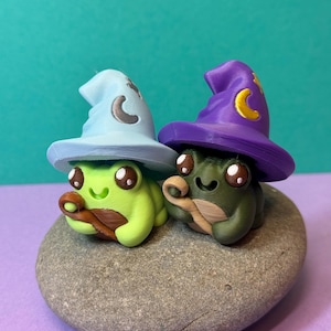May include: Two whimsical frog figurines, one green and one olive, each wearing a wizard hat. The hats are light blue and purple, adorned with crescent moon details. The frogs sit on a gray stone, with a teal background.