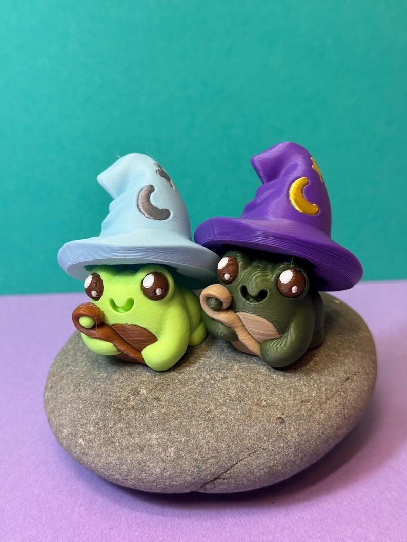 May include: Two whimsical frog figurines, one green and one olive, each wearing a wizard hat. The hats are light blue and purple, adorned with crescent moon details. The frogs sit on a gray stone, with a teal background.