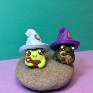 May include: Two whimsical frog figurines, one light green and the other dark green, each wearing a wizard hat. The light green frog has a light blue hat with a crescent moon, while the dark green frog has a purple hat with a yellow circle. They sit on a gray stone.