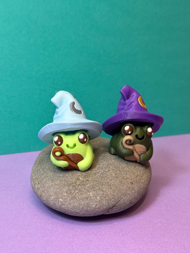 May include: Two whimsical frog figurines, one light green and the other dark green, each wearing a wizard hat. The light green frog has a light blue hat with a crescent moon, while the dark green frog has a purple hat with a yellow circle. They sit on a gray stone.