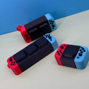 May include: Three black, red, and blue game controllers are displayed on a light surface against a blue backdrop. The controllers have a rectangular black centre section with red and blue ends, featuring buttons and control pads.
