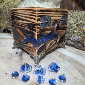 May include: A square wooden box with a scorched finish, featuring ornate metal hinges and decorative corner feet. The front panel displays a blue resin inlay with a wing-like design. Eight clear, multi-faceted polyhedral dice with blue inclusions are arranged in front of the box.
