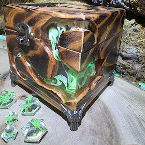 May include: A square wooden box featuring dark scorched wood grain and integrated swirls of translucent green and white resin. The box has a silver-toned metal latch and decorative corner feet, accompanied by five small, faceted green resin gems.