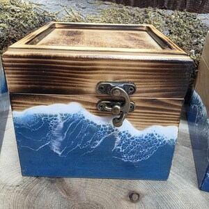Op de afbeelding: Een vierkante houten doos met een scharnierend deksel en een metalen sluiting. Het bovenste gedeelte van de doos heeft een natuurlijke gebrande houtafwerking, terwijl het onderste gedeelte is versierd met een blauw en wit harsontwerp.