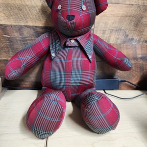 May include: A handmade teddy bear made of red plaid fabric with blue and green accents. The bear has button eyes and a black nose. It is wearing a collared shirt with button details. A unique, handcrafted plush toy.