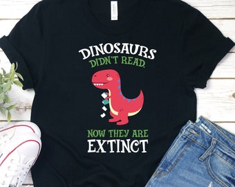May include: Black t-shirt with a green and white graphic of a dinosaur holding a stack of books. The text "Dinosaurs didn't read. Now they are extinct" is printed on the shirt.