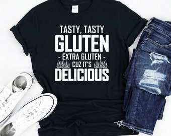 Puede incluir: Camiseta negra con texto blanco que dice "Tasty, Tasty Gluten - Extra Gluten - Cuz It's Delicious". El texto está rodeado de tallos de trigo.