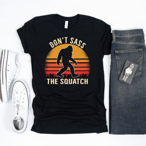 May include: Black t-shirt with a retro sunset graphic featuring a silhouette of Bigfoot walking. The text "Don't Sass The Squatch" is printed in white.