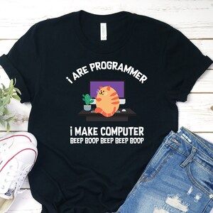 Pode incluir: Camiseta preta com um gráfico branco de um gato sentado em frente a um computador. O texto na camiseta diz "I are programmer I make computer beep boop beep beep boop."
