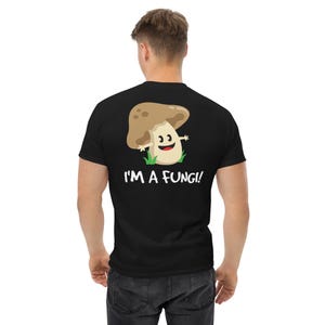 May include: A black short-sleeved t-shirt featuring a cartoon mushroom graphic on the back. The mushroom has a tan cap, a cream-colored stem, and a smiling face. Below the graphic, the white text reads, I'M A FUNGI! with green grass accents.