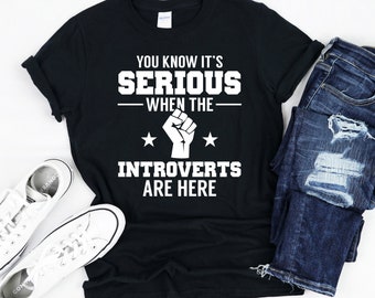 Może przedstawiać: Czarny T-shirt z białym tekstem, który brzmi "You know it's serious when the introverts are here". Tekst jest otoczony dwiema białymi gwiazdami. W środku tekstu znajduje się biała pięść.