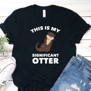 Könnte beinhalten: Schwarzes T-Shirt mit weißem Text, der "This is my significant otter" lautet. Ein Cartoon-Otter hält eine Blume im Maul.