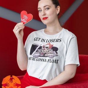 May include: A white t-shirt featuring a graphic of Pennywise the clown from the film "IT" driving a car. The text "Get in Losers, We're Gonna Float" is printed on the shirt. A person is holding a red heart-shaped cookie that says "You Wish".