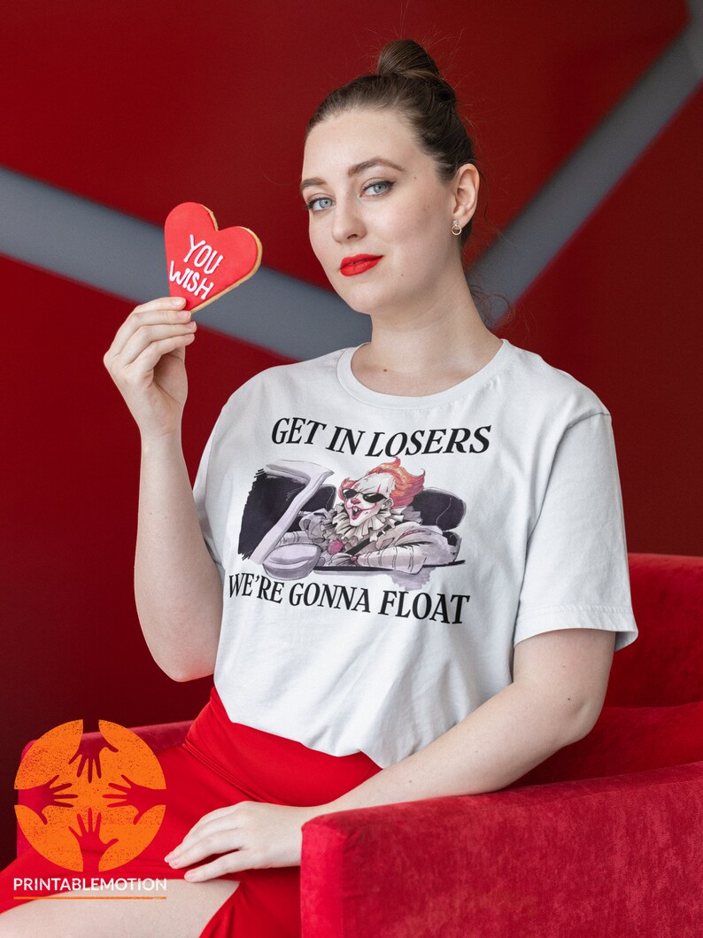 May include: A white t-shirt featuring a graphic of Pennywise the clown from the film "IT" driving a car. The text "Get in Losers, We're Gonna Float" is printed on the shirt. A person is holding a red heart-shaped cookie that says "You Wish".