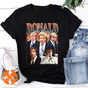 Puede incluir: Camiseta negra con un gráfico de estilo vintage de una banda llamada "DONALD John Trump". El gráfico presenta a cuatro hombres con una estética retro de los años 80.