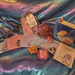 May include: A collection of six tea bags featuring Alice in Wonderland illustrations. The tea bags are packaged in a white and blue box with the words "Wonders of Tea" printed on the front. The tea bags are arranged on a blue and purple iridescent fabric background.