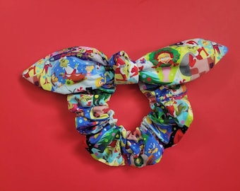 May include: A festive Christmas scrunchie with a bow, featuring a vibrant print of cartoon characters and holiday motifs. The scrunchie is designed for hair styling and is set against a red backdrop.