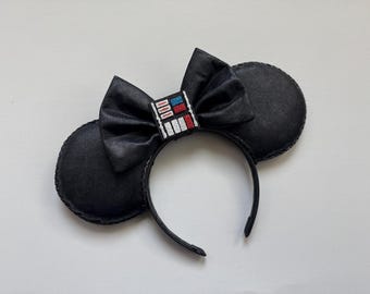 May include: Black mouse ear headband with a satin bow and two round ears. The bow features a rectangular design with red, white, and blue accents. The headband is a fun accessory for themed events.