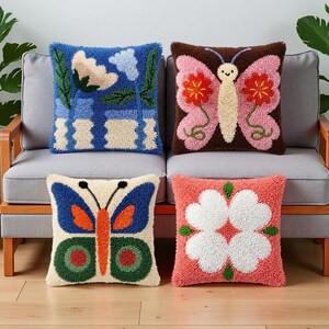 Op de afbeelding: Vier decoratieve kussens met kleurrijke, getextureerde ontwerpen. Eén kussen heeft een blauw bloemenpatroon, een ander een vlinder, en twee andere tonen vlinder- en bloemmotieven. De kussens liggen op een grijze bank.