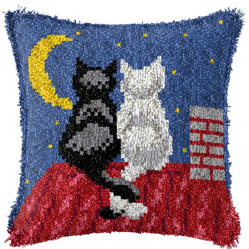 以下が含まれることがあります： 青い背景に、赤い屋根の上に座っている2匹の猫が描かれた、ぬいぐるみ製のクッション。猫の左側は灰色と黒で、右側は白です。猫たちは、黄色い三日月と星の下にいます。