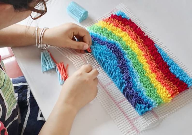 Op de afbeelding: Een kleurrijk regenboogkleed wordt gemaakt met een naald en garen. Het kleed heeft banden in rood, oranje, geel, groen, blauw en paars. Het werk bevindt zich op een witte mesh-basis, met blauwe en rode kwasten in de buurt.