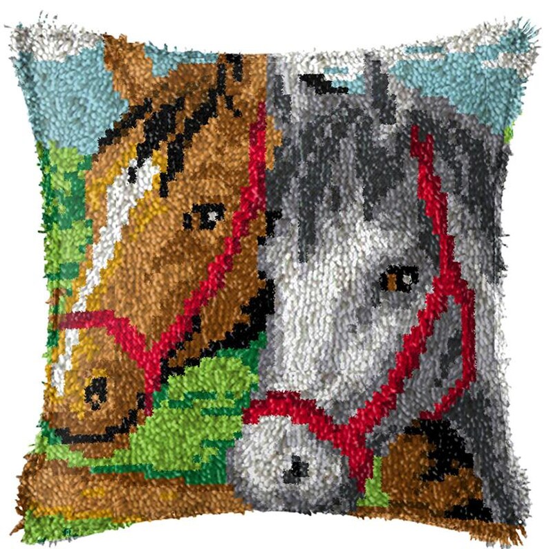 以下が含まれることがあります： 2頭の馬の顔がアップで描かれたカラフルな枕。馬は茶色と灰色で、赤い手綱をしています。背景は緑の草原と青い空です。