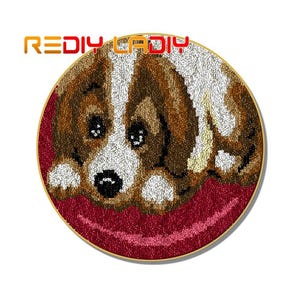 May include: A close-up of a brown and white dog with brown eyes, lying on a red background. The dog is made of yarn and is a rug hooking project.