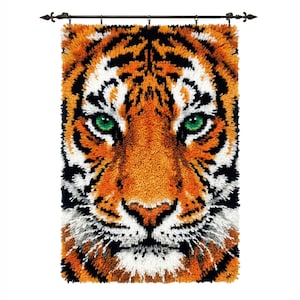 Könnte beinhalten: Ein Wandbehang mit einem Tigergesicht im Nahaufnahme. Der Tiger hat orangefarbenes Fell mit schwarzen Streifen, weißen Akzenten und auffälligen grünen Augen. Montiert an einer Stange mit dekorativen Endstücken.