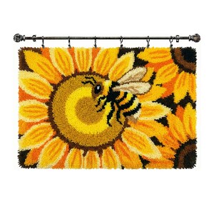 May include: A rectangular latch hook wall hanging featuring a large yellow sunflower with a brown center and a black and yellow bumblebee resting on the petals. The textile piece is suspended from a dark metal rod with six silver hanging rings.