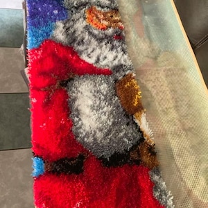 Op de afbeelding: Een gedeeltelijk afgemaakt Santa Claus-tapijt, met een rood pak en hoed, witte baard en een blauwe en paarse sterrenhemel achtergrond. Het tapijt is gemaakt met een getextureerd, gelust garen en is gedeeltelijk bevestigd aan een gaasrug.