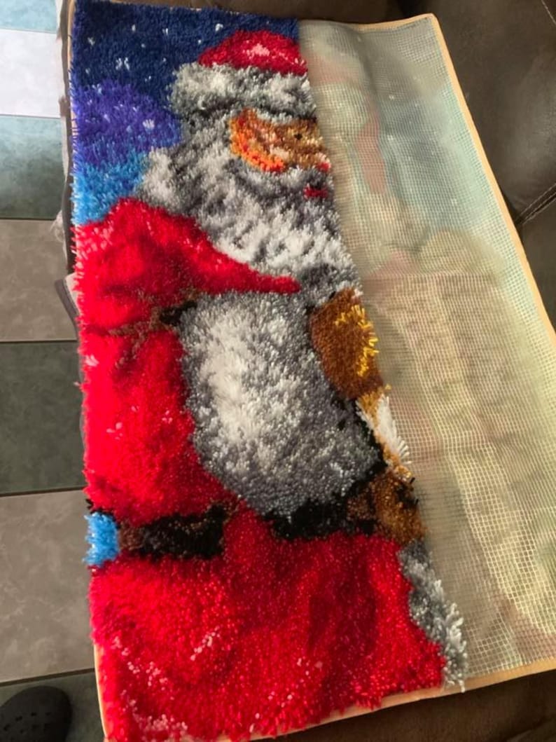 Op de afbeelding: Een gedeeltelijk afgemaakt Santa Claus-tapijt, met een rood pak en hoed, witte baard en een blauwe en paarse sterrenhemel achtergrond. Het tapijt is gemaakt met een getextureerd, gelust garen en is gedeeltelijk bevestigd aan een gaasrug.