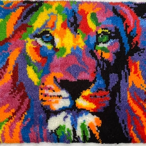 Op de afbeelding: Een kleurrijk, handgemaakt tapijt met een leeuwenkop. De manen en het gezicht van de leeuw zijn afgebeeld in een levendig kleurenpalet, waaronder blauw, oranje, geel en roze, tegen een zwarte achtergrond. Het tapijt is gemaakt van garen.
