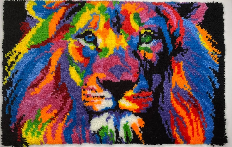 Op de afbeelding: Een kleurrijk, handgemaakt tapijt met een leeuwenkop. De manen en het gezicht van de leeuw zijn afgebeeld in een levendig kleurenpalet, waaronder blauw, oranje, geel en roze, tegen een zwarte achtergrond. Het tapijt is gemaakt van garen.