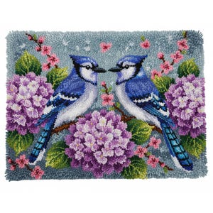 May include: A hand-stitched rug featuring two blue jays perched on a branch, facing each other. The birds are surrounded by purple hydrangeas, pink blossoms, and green leaves against a light blue background. The rug has a textured, handmade appearance.
