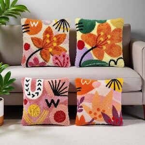 Puede incluir: Cuatro cojines cuadrados con diseños florales y abstractos coloridos. Los cojines presentan tonos naranja, rosa, verde y amarillo. Cada cojín tiene un patrón único, que incluye flores, formas y la letra "W".