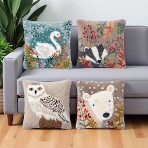 May include: Four square throw pillows featuring textured punch needle embroidery. The designs include a white swan on a teal background, a badger among autumn foliage, a snowy owl on a branch, and a polar bear face surrounded by colorful plants.