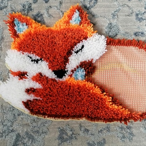 Op de afbeelding: Handgemaakt vosvormig tapijt in oranje, wit en zwart, met blauwe accenten op de oren. Het tapijt heeft een pluizig, getextureerd uiterlijk. Het ligt op een patroon, lichtblauw en grijs oppervlak.