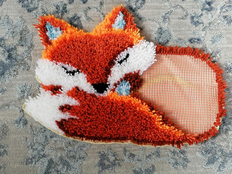 Op de afbeelding: Handgemaakt vosvormig tapijt in oranje, wit en zwart, met blauwe accenten op de oren. Het tapijt heeft een pluizig, getextureerd uiterlijk. Het ligt op een patroon, lichtblauw en grijs oppervlak.