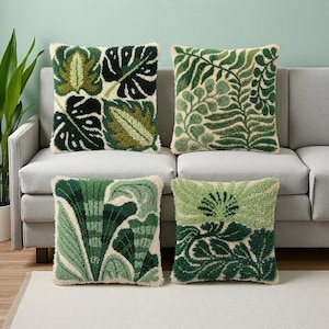 May include: Four square throw pillows with botanical designs. Each pillow features a different green leaf pattern, including monstera leaves and ferns, on a cream background. The pillows are arranged on a gray sofa, creating a natural, decorative theme.