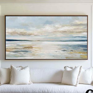 Puede incluir: Pintura abstracta de paisaje rectangular con marco dorado. La obra presenta una escena serena con cielo azul claro, nubes y agua en tonos beige y azul. Debajo, un sofá blanco con cojines.