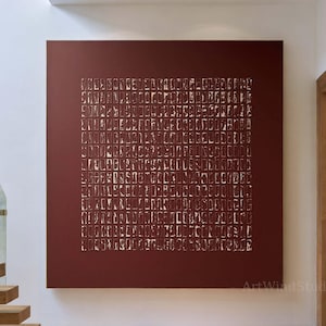 May include: A large square wall art piece featuring a deep burgundy background with a grid of numerous small, vertical rectangular shapes in a metallic gold finish. The artwork is displayed on a white wall in a hallway with wooden stairs nearby.