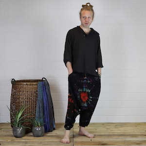 May include: A person wearing a black hooded shirt and tie-dye pants. The pants have a large, colorful swirl design. The person is standing in front of a white brick wall.
