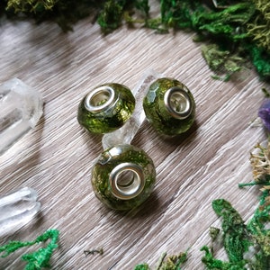 May include: Three green and silver charm beads with a moss-like interior. The beads are round with a faceted surface and a silver-colored metal core. The beads are on a light wood surface with green moss and crystals.