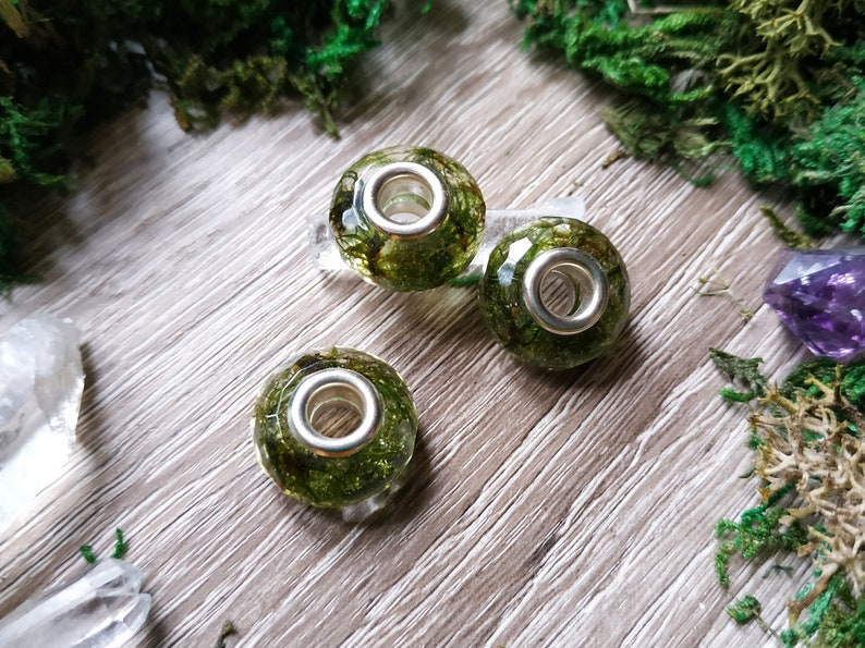May include: Three round beads with a silver-colored core, filled with green moss. The beads are clear, allowing the moss to be visible. The beads are on a wooden surface, surrounded by green moss and crystals.
