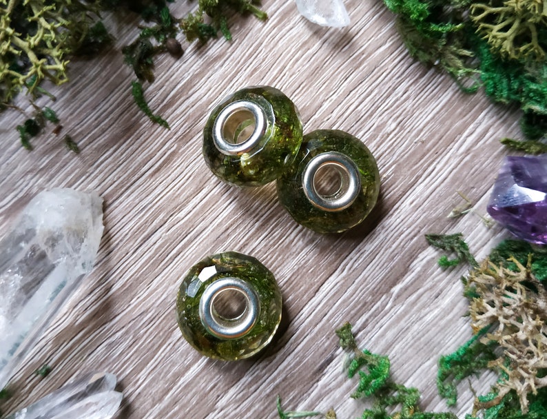 May include: Three clear glass beads filled with green moss and small silver-colored metal rings. The beads are on a wooden surface with green moss and crystals.