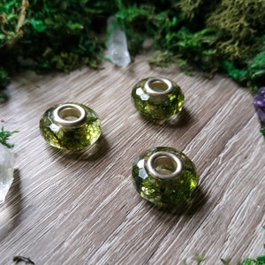 May include: Three faceted glass beads with a silver-colored core, filled with green plant material. The beads are arranged on a wooden surface, surrounded by green moss, crystals, and a purple gemstone.