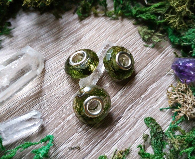 May include: Three green and silver charm beads with a moss-like interior. The beads are round with a faceted surface and a silver-colored metal core. The beads are on a light wood surface with green moss and crystals.