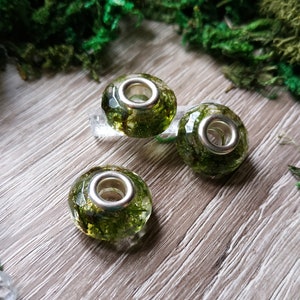 May include: Three clear, faceted glass beads with silver-colored metal cores, each encapsulating green moss. The beads are displayed on a light wood surface, surrounded by green moss and crystals, creating a natural, earthy aesthetic.
