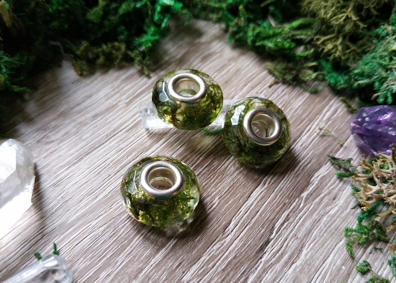 May include: Three clear, faceted glass beads with silver-colored metal cores, each encapsulating green moss. The beads are displayed on a light wood surface, surrounded by green moss and crystals, creating a natural, earthy aesthetic.